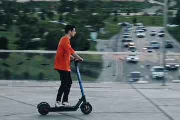 a guy in an orange T-shirt rides an electric scooter over a bridge